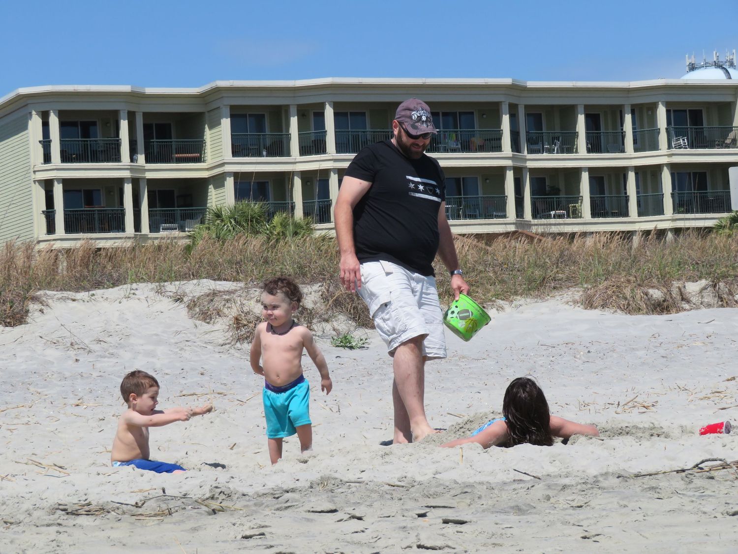 A man with three young children playing in the sand near a beachfront hotel.