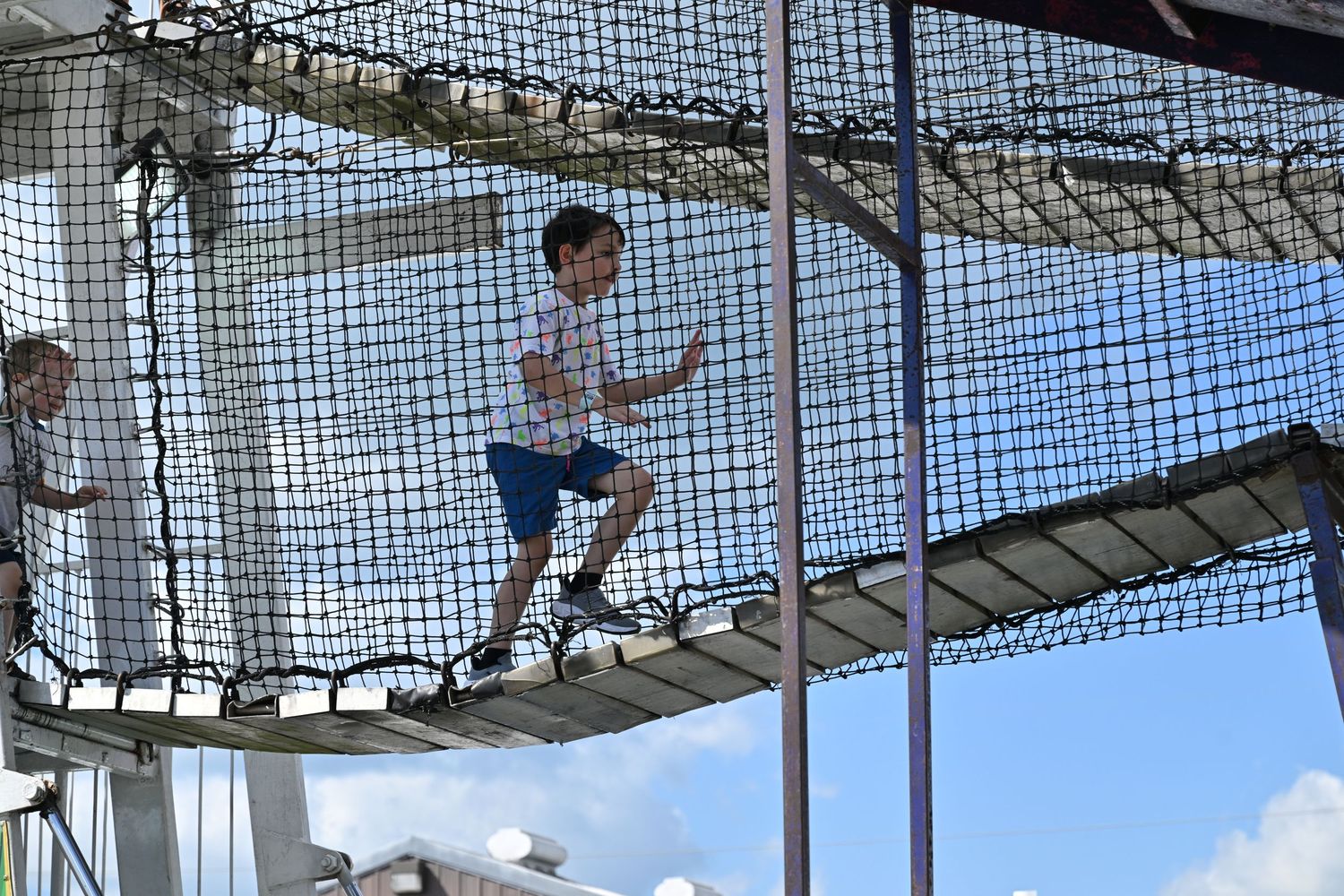 A young boy walks across a rope bridge at an outdoor adventure playground on a sunny day.