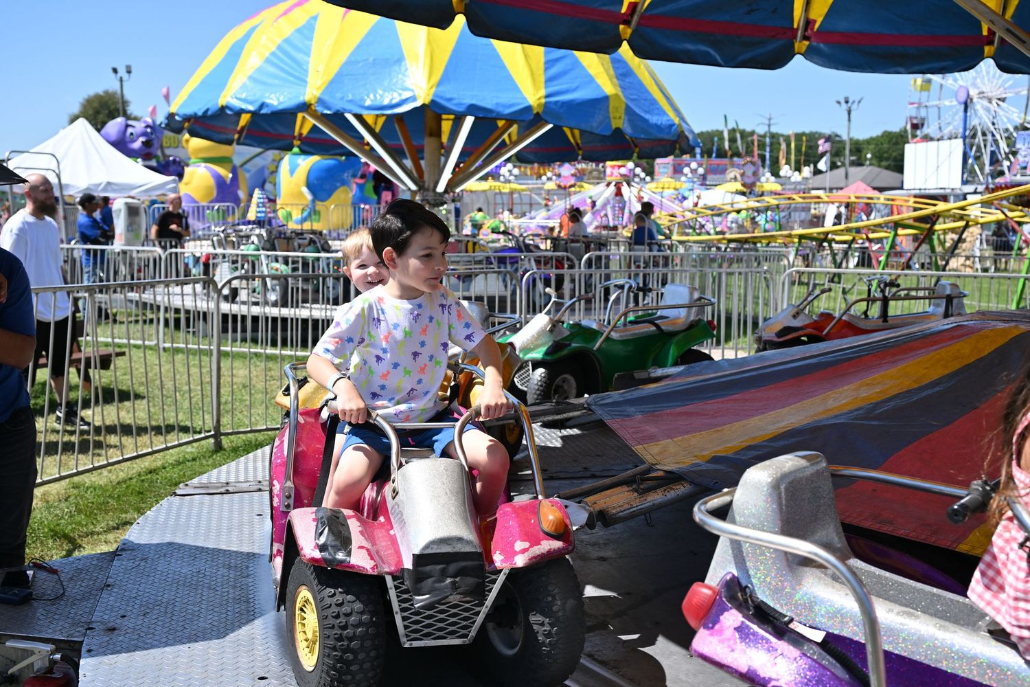 Young child rides a pink carnival car with stuffed animals at a sunny fairground.
