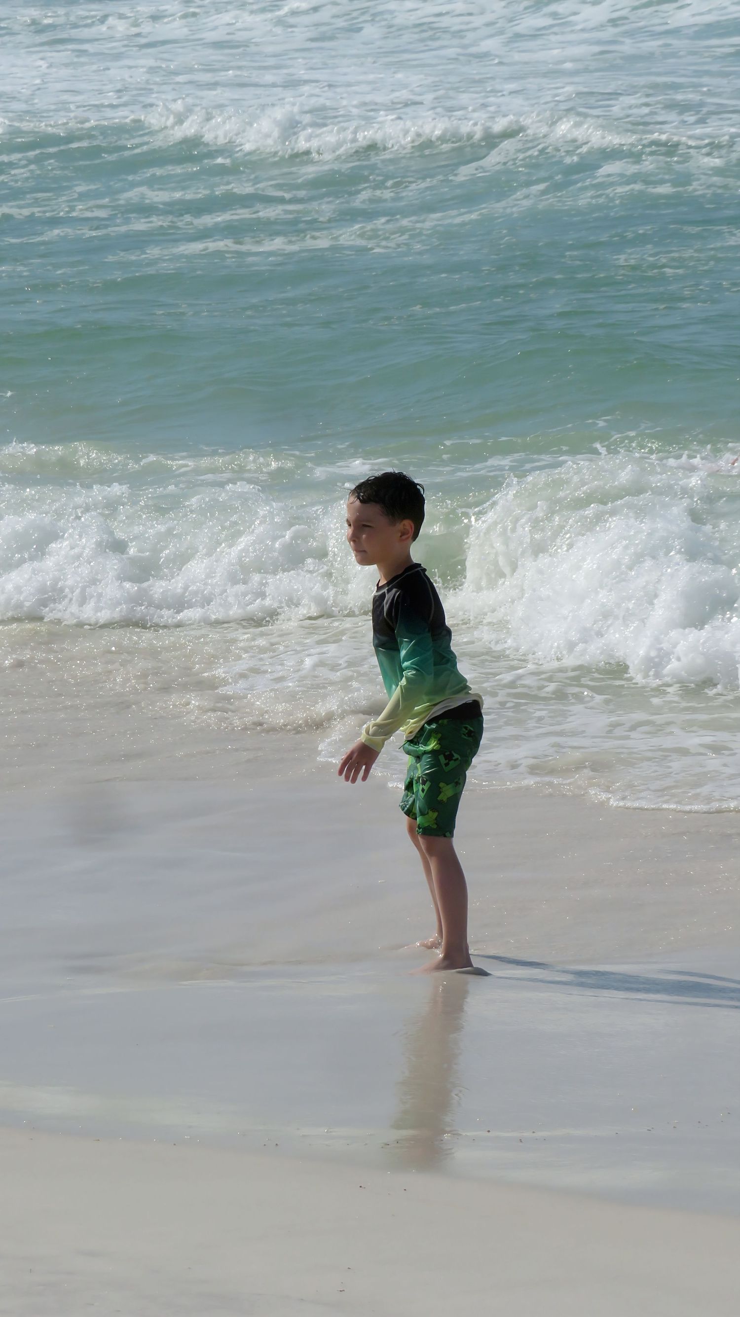 Boy in green rash guard stands at water's edge on white sandy beach with turquoise waves.