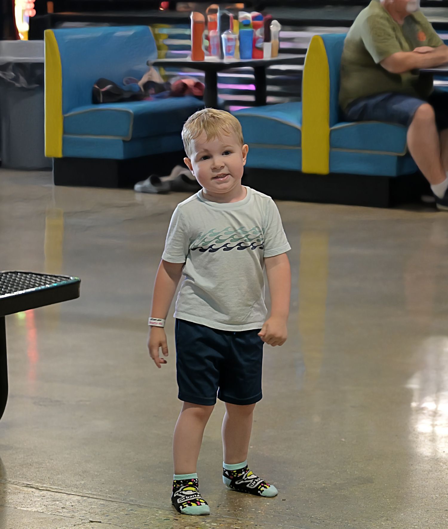 A toddler in a white shirt and navy shorts stands confidently at an indoor bowling alley.