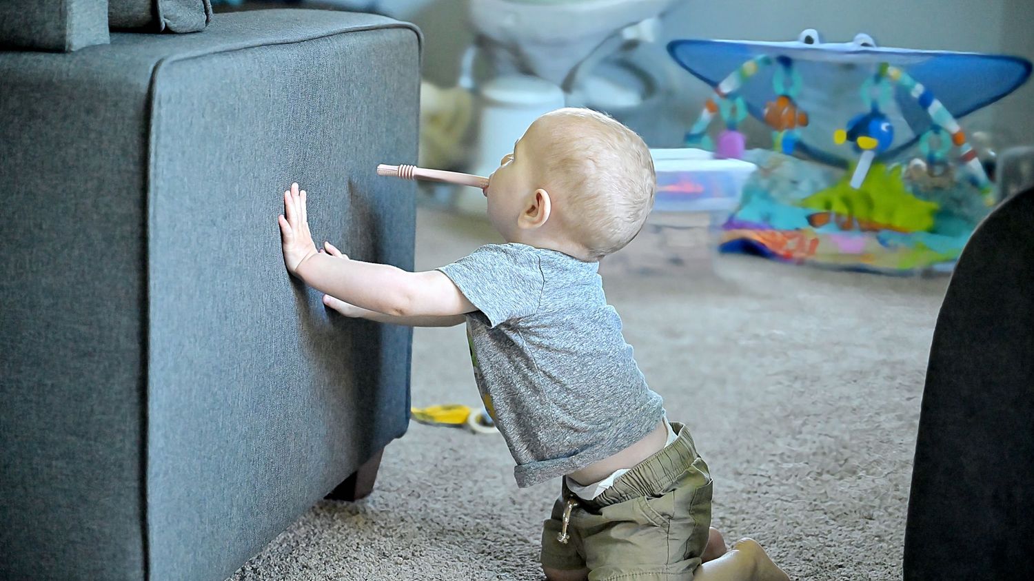 A baby in a gray shirt practices standing by pressing hands against the side of a sofa.