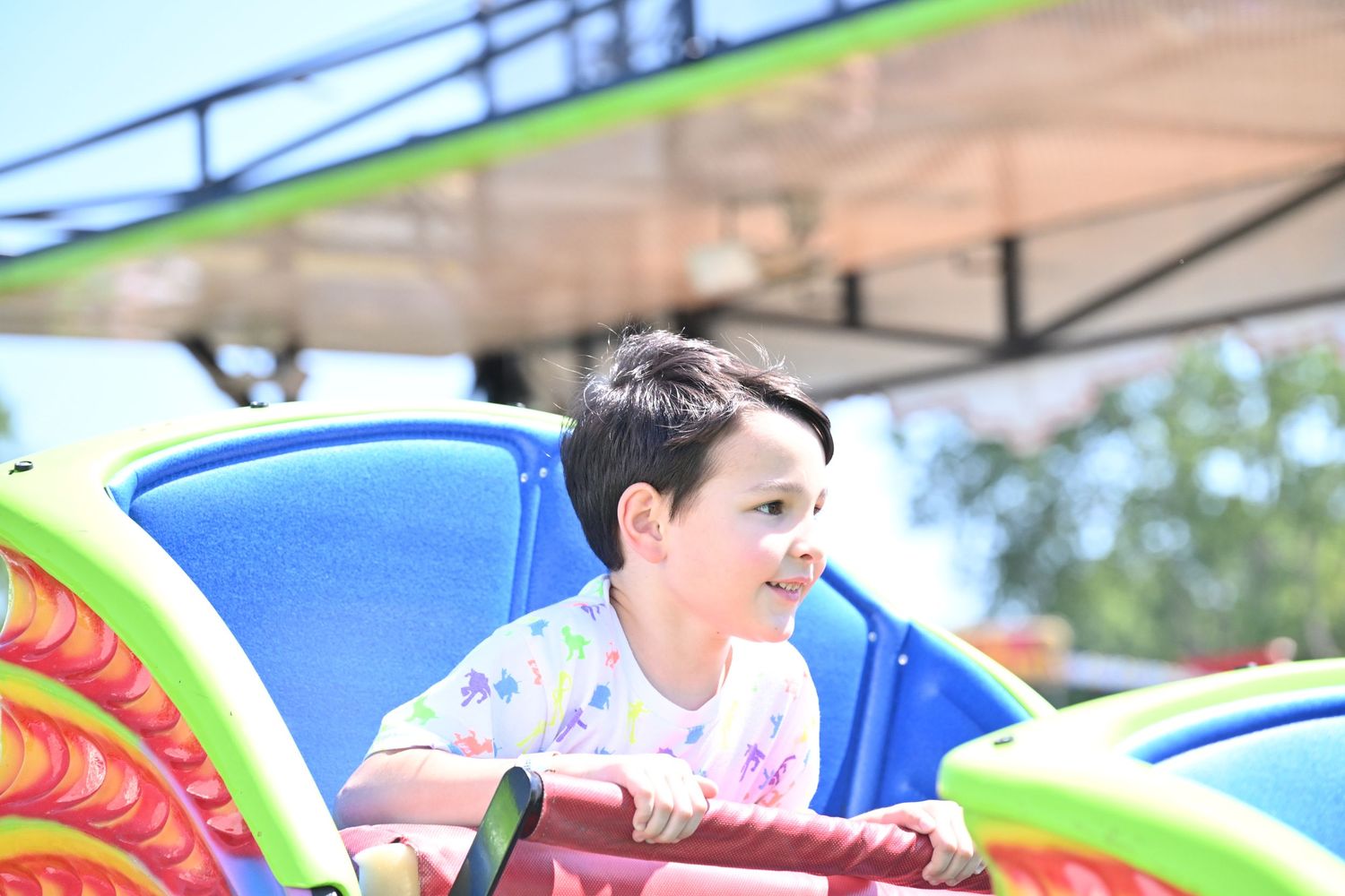 Young boy smiles while riding a colorful carnival ride on a sunny day.