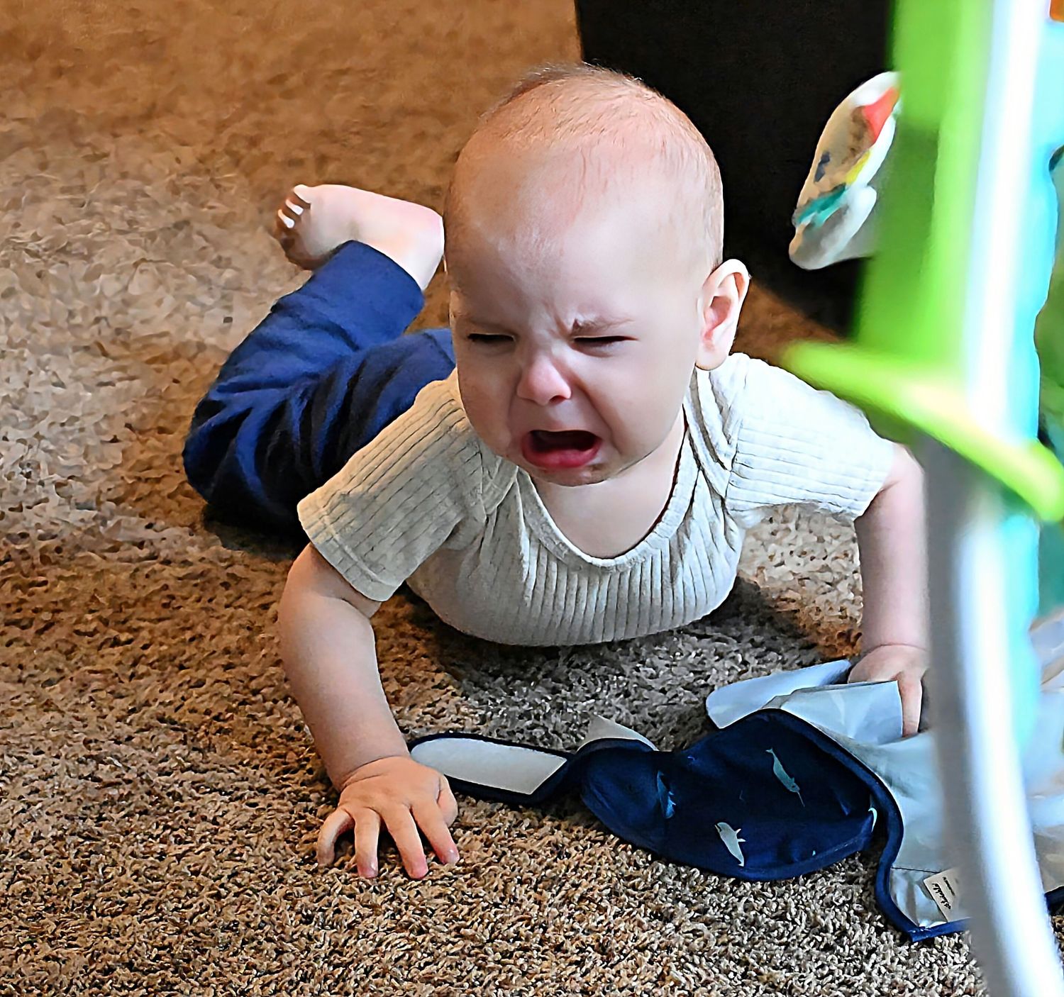 A crying baby in a white onesie lies on a beige carpet next to colorful toys.