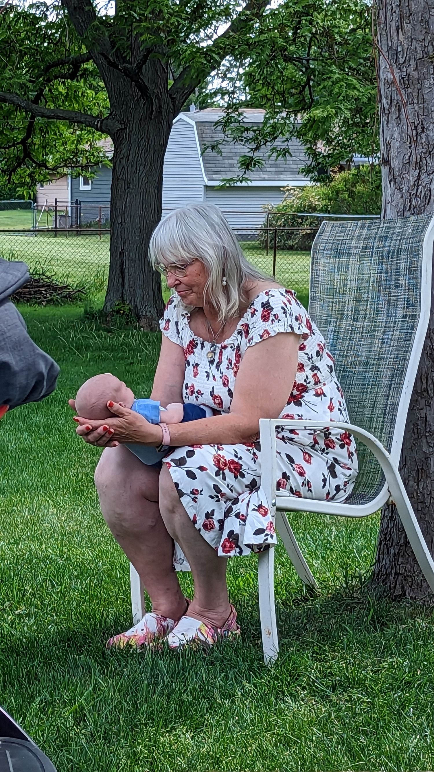 Smiling grandmother in floral dress sits outdoors on chair holding newborn baby under tree.