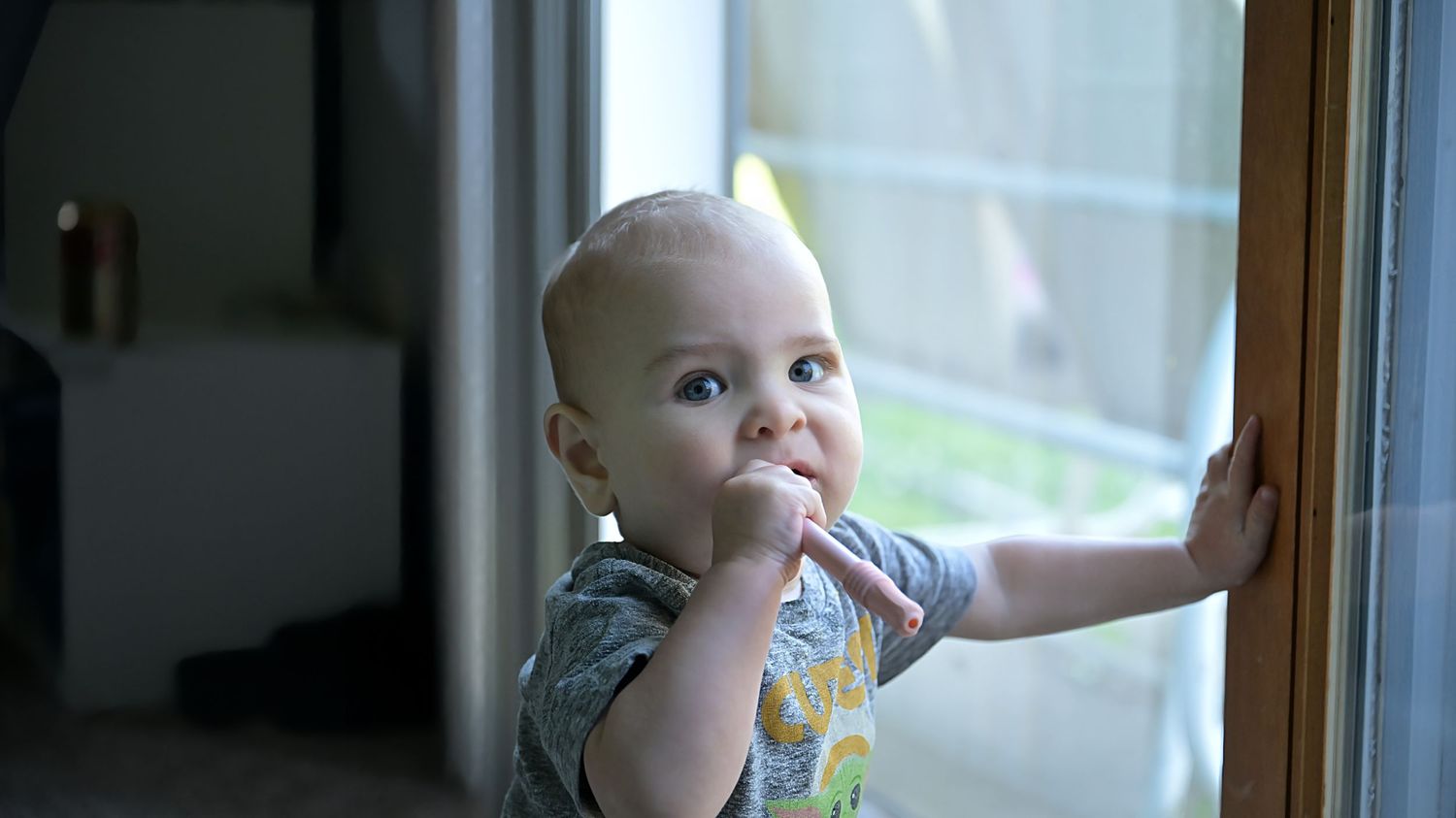 A baby stands at a glass sliding door, fingers in mouth, gazing outside with bright eyes.