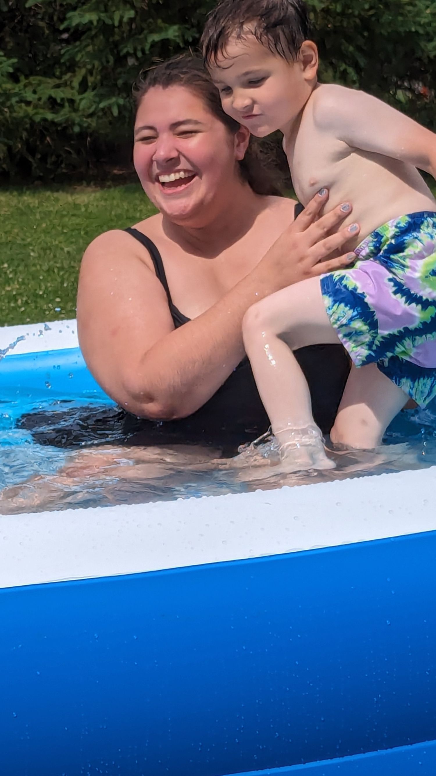 A laughing woman holds a young boy in a blue inflatable backyard pool on a summer day.