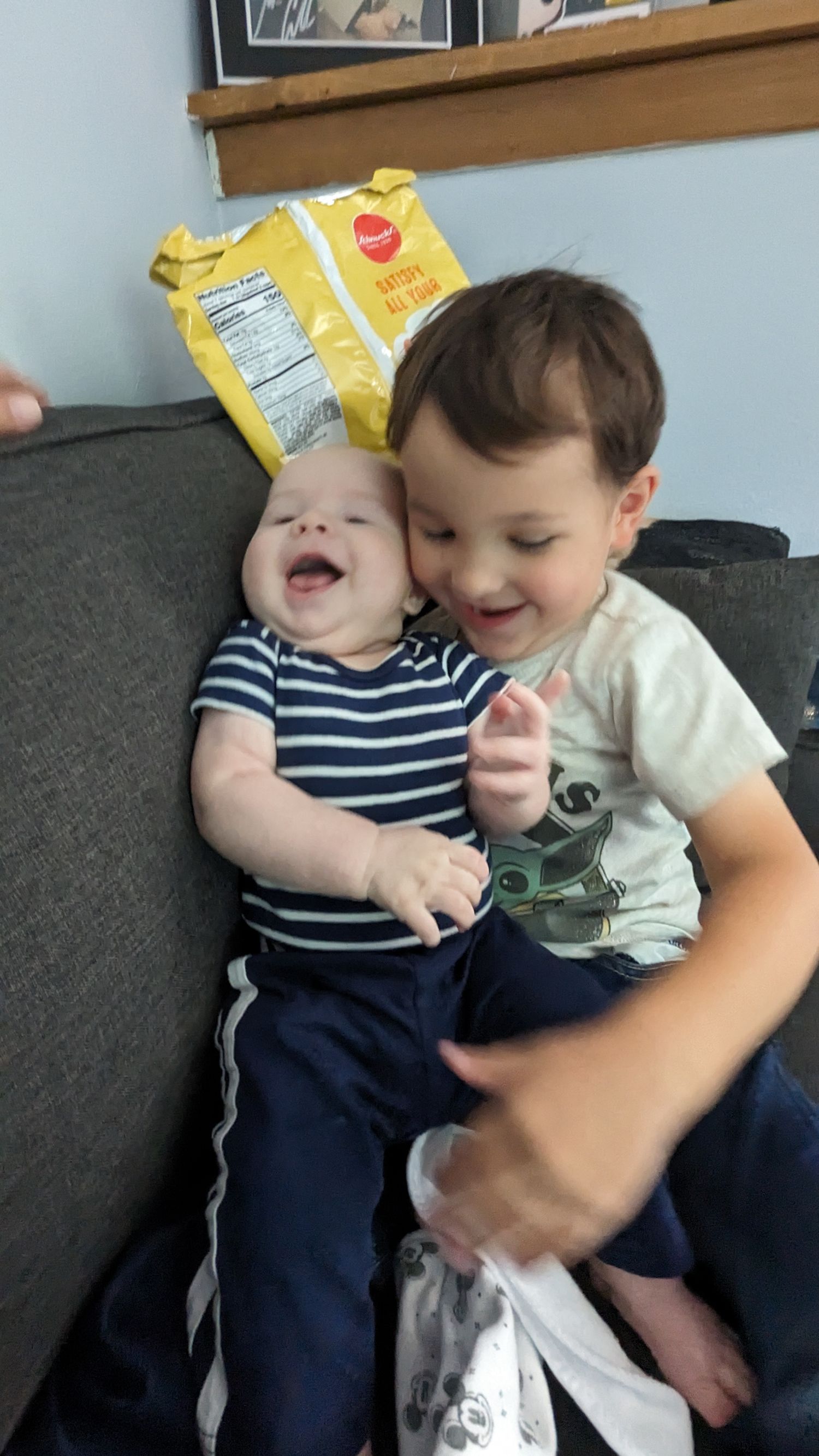 Happy toddler hugs laughing baby sibling on gray couch at home.