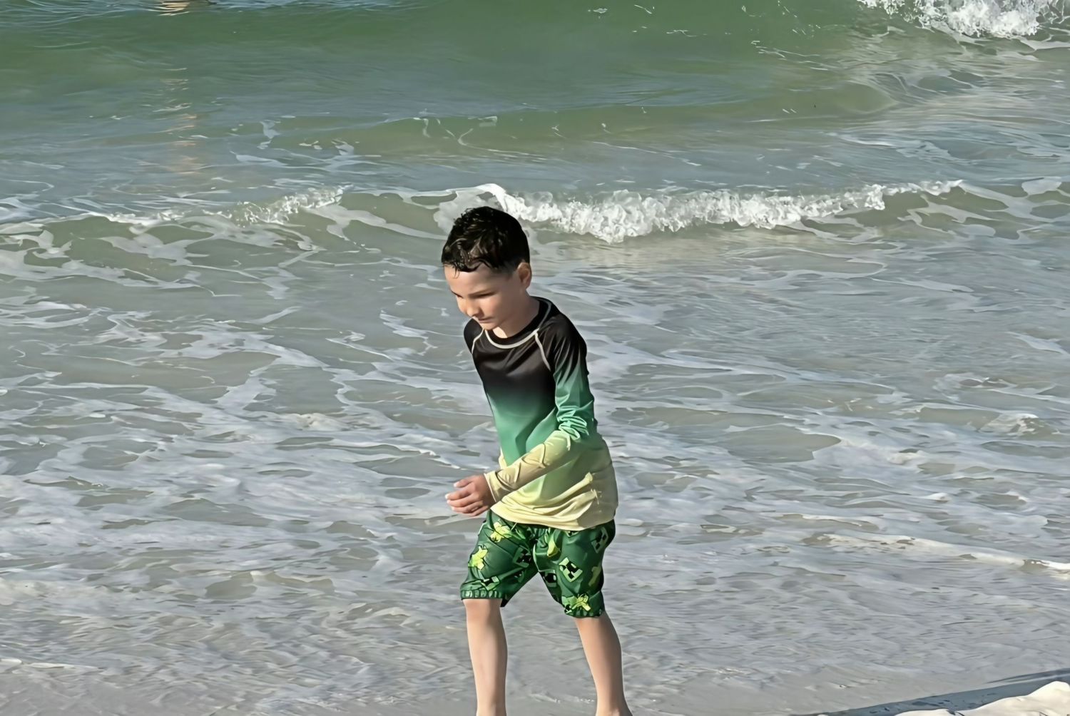 A boy in green rash guard and swim trunks plays at the ocean shoreline with waves behind him.
