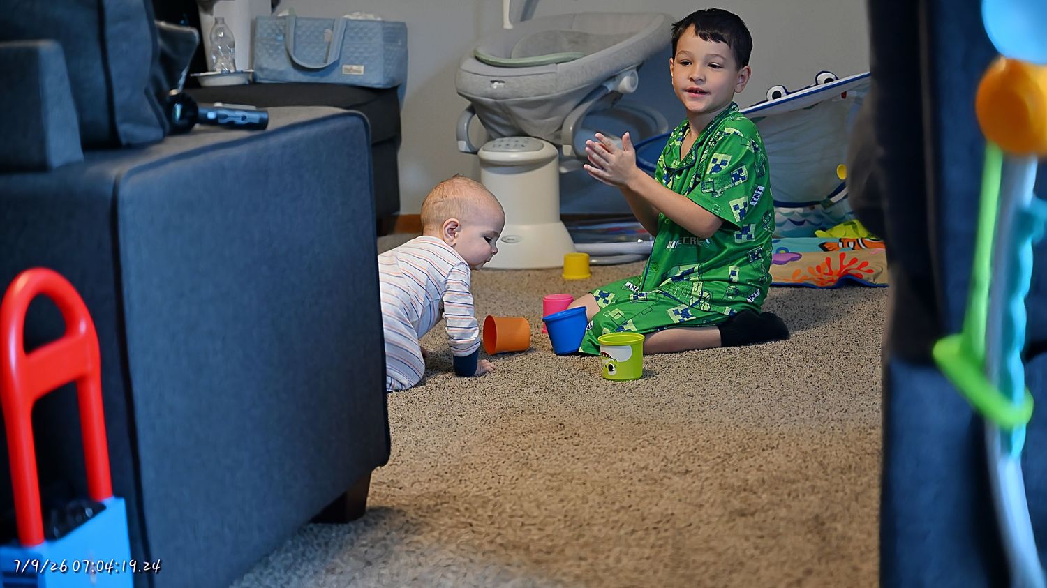A boy in green pajamas clapping and playing with a crawling baby on a carpeted floor.