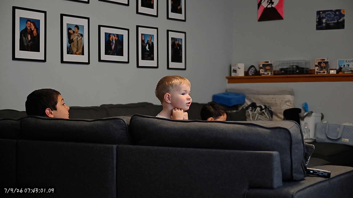 Two children sit on a gray sofa watching TV in a living room decorated with framed photos.