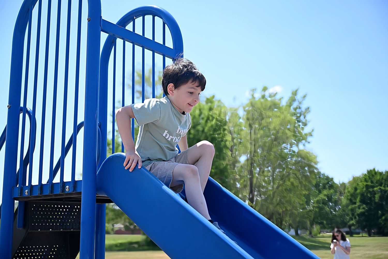 A young boy in a green shirt sits at the top of a blue playground slide on a sunny day.