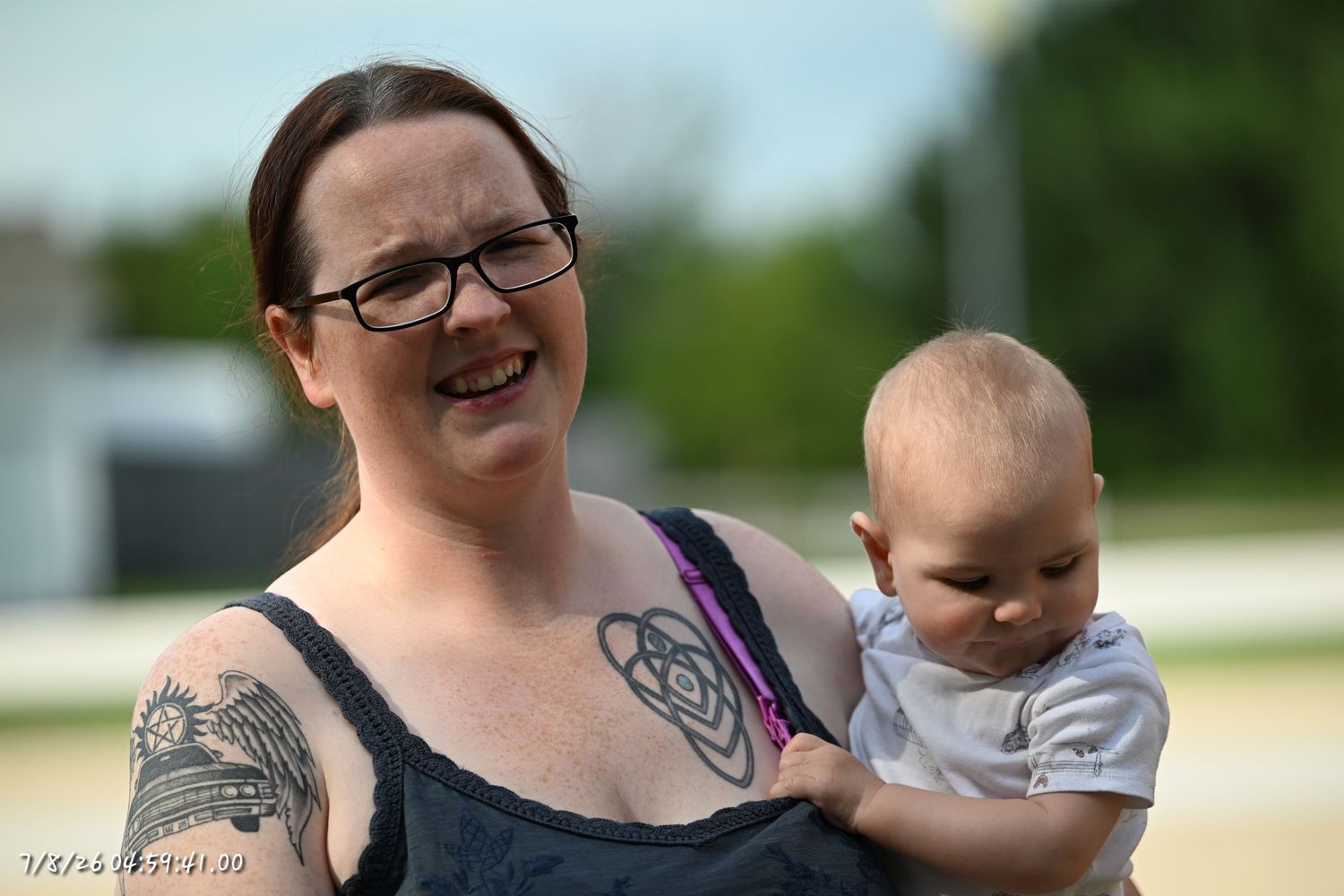Tattooed mother holding her young baby outdoors on a sunny day.