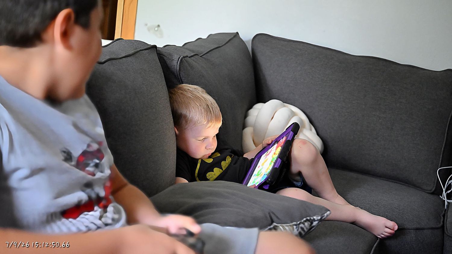 Two boys sit on a gray couch, one using a tablet device in a cozy home setting.