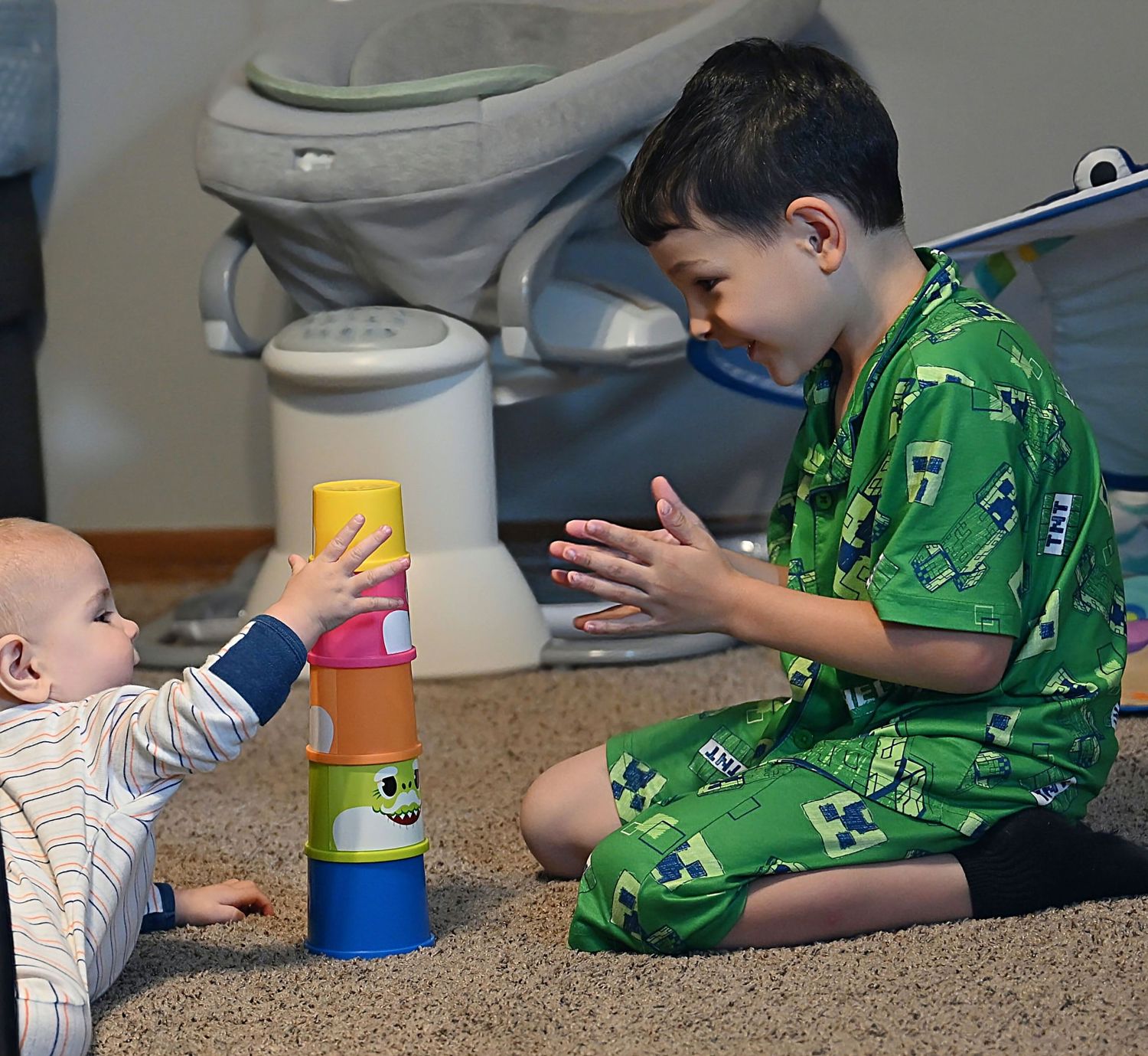 A young boy in green pajamas plays stacking cups with a smiling baby on a carpeted floor.