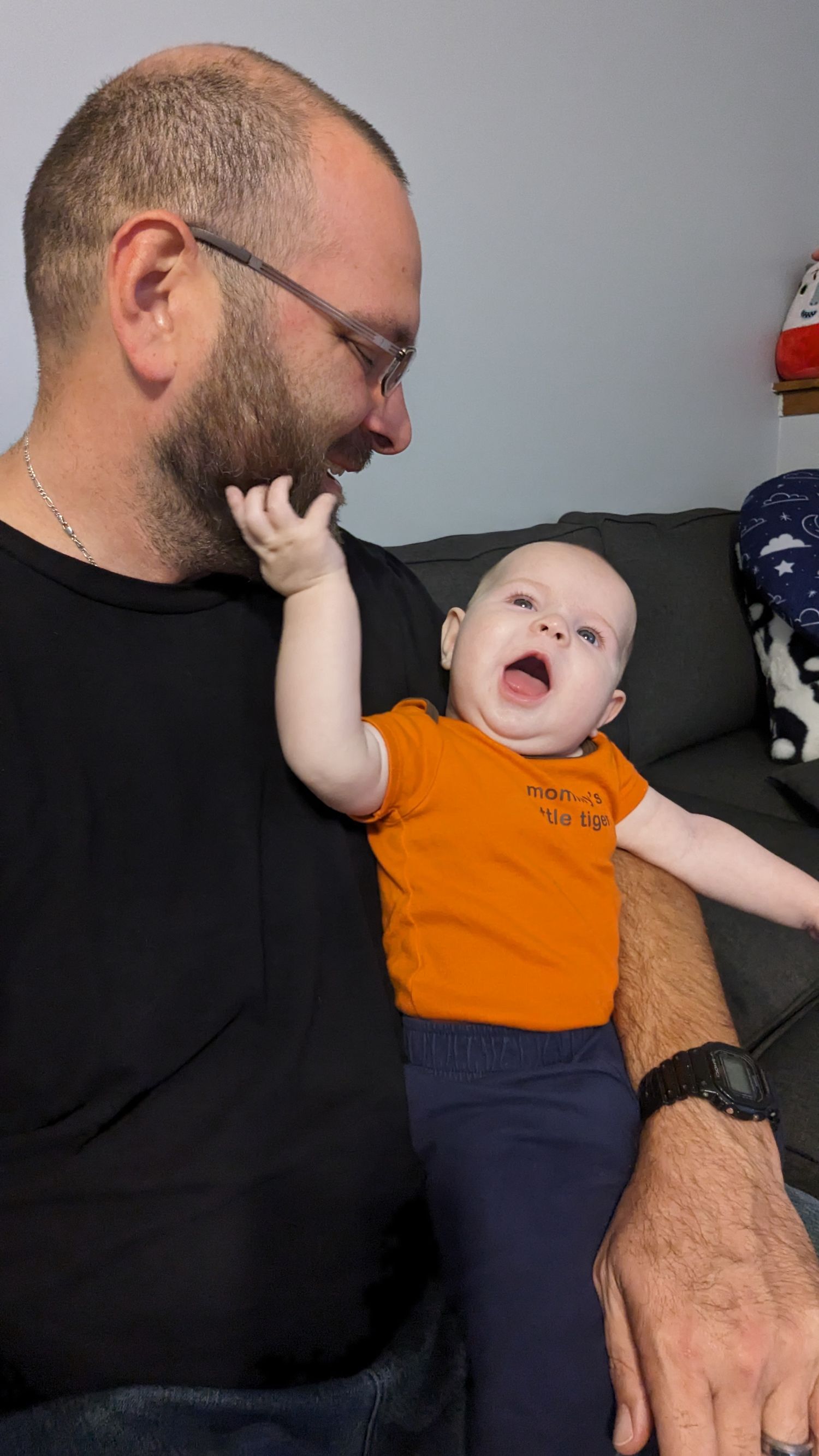 A bearded father in a black shirt holds his laughing baby in an orange onesie indoors.
