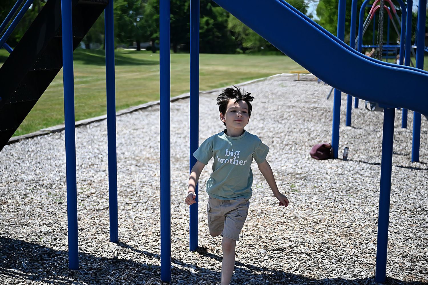 A smiling boy wearing a big brother shirt runs through a blue playground structure on a sunny day.