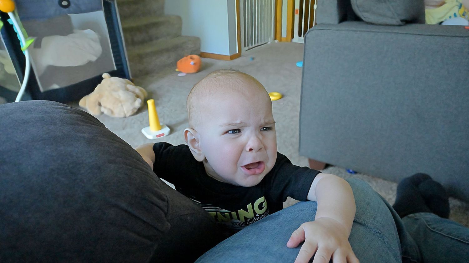 An expressive baby in a black shirt makes a funny face while leaning on a gray couch.