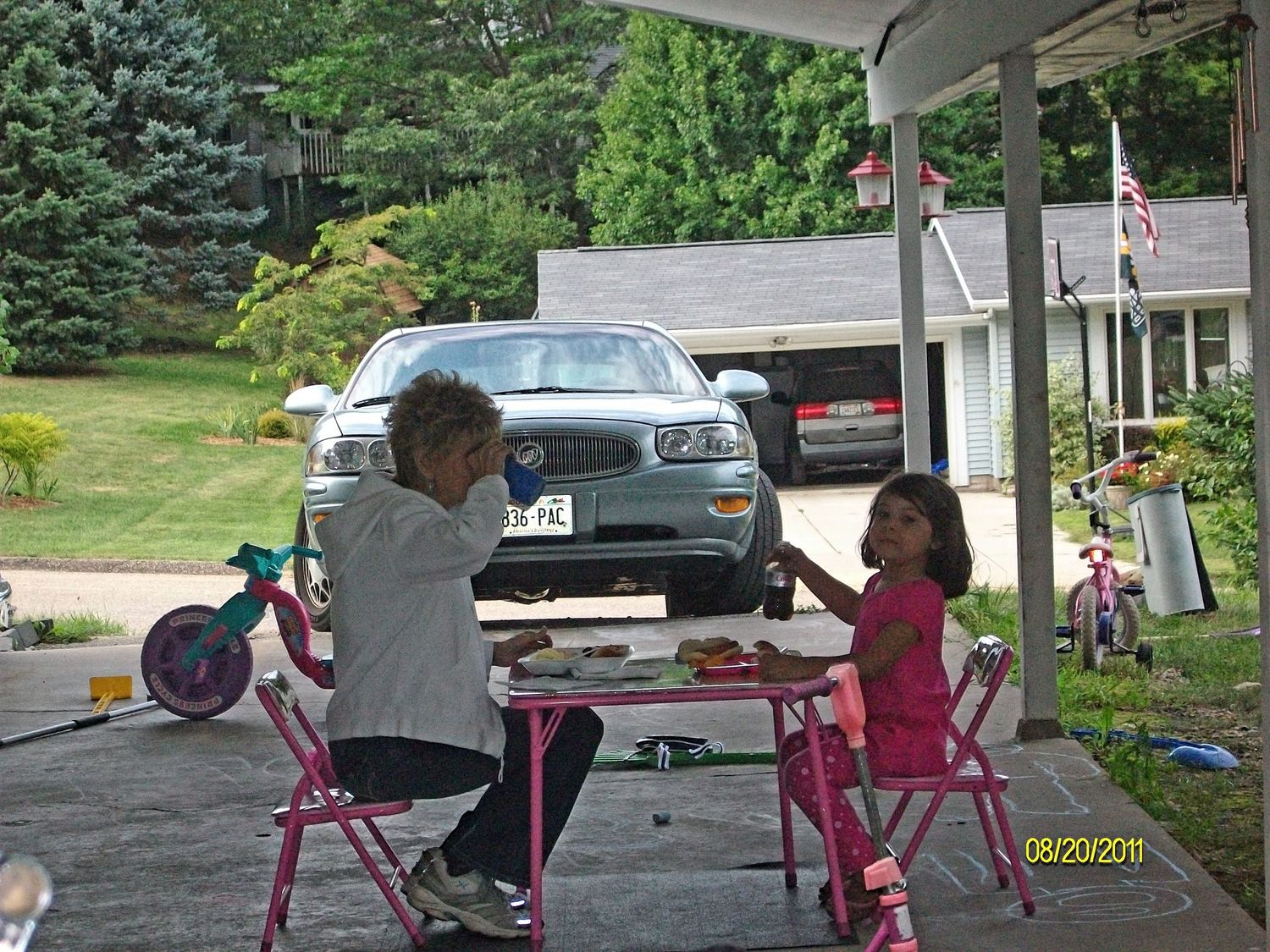 Nana sitting at a pink folding table on a driveway, eating with granddaughter outdoors near a parked car on a sunny day.