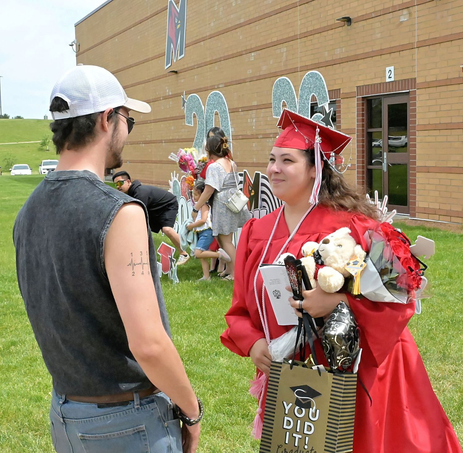 Female graduate in red cap and gown holds flowers and gifts while chatting with a guest outside school.