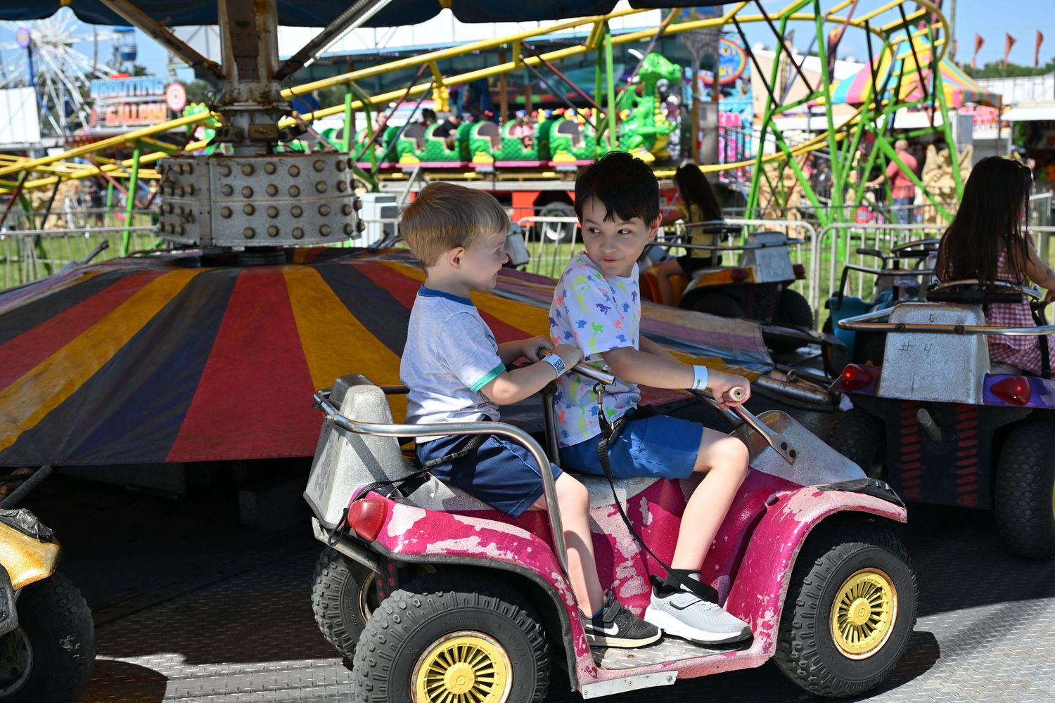 Two children ride a colorful carnival car ride at a sunny outdoor fair.