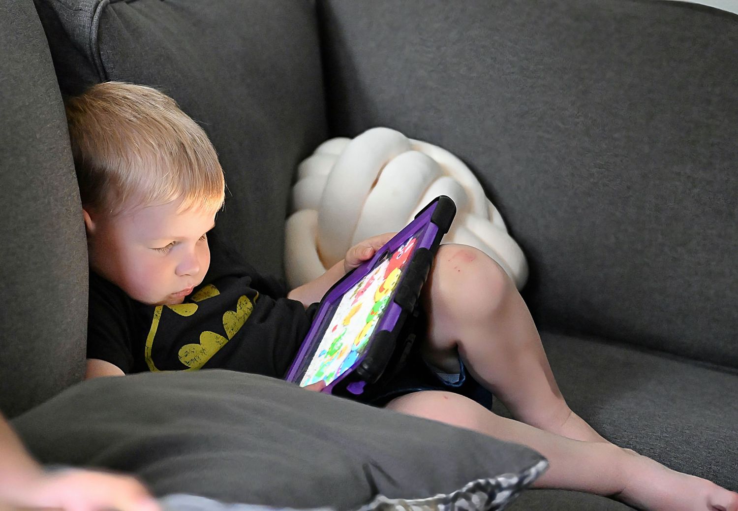 A young boy in a Batman shirt falls asleep watching a tablet on a gray couch with pillows.