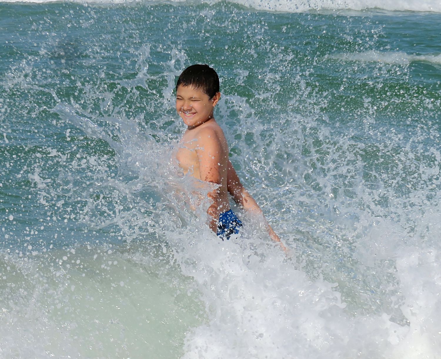 Happy boy laughs as ocean waves crash around him while swimming at the beach.