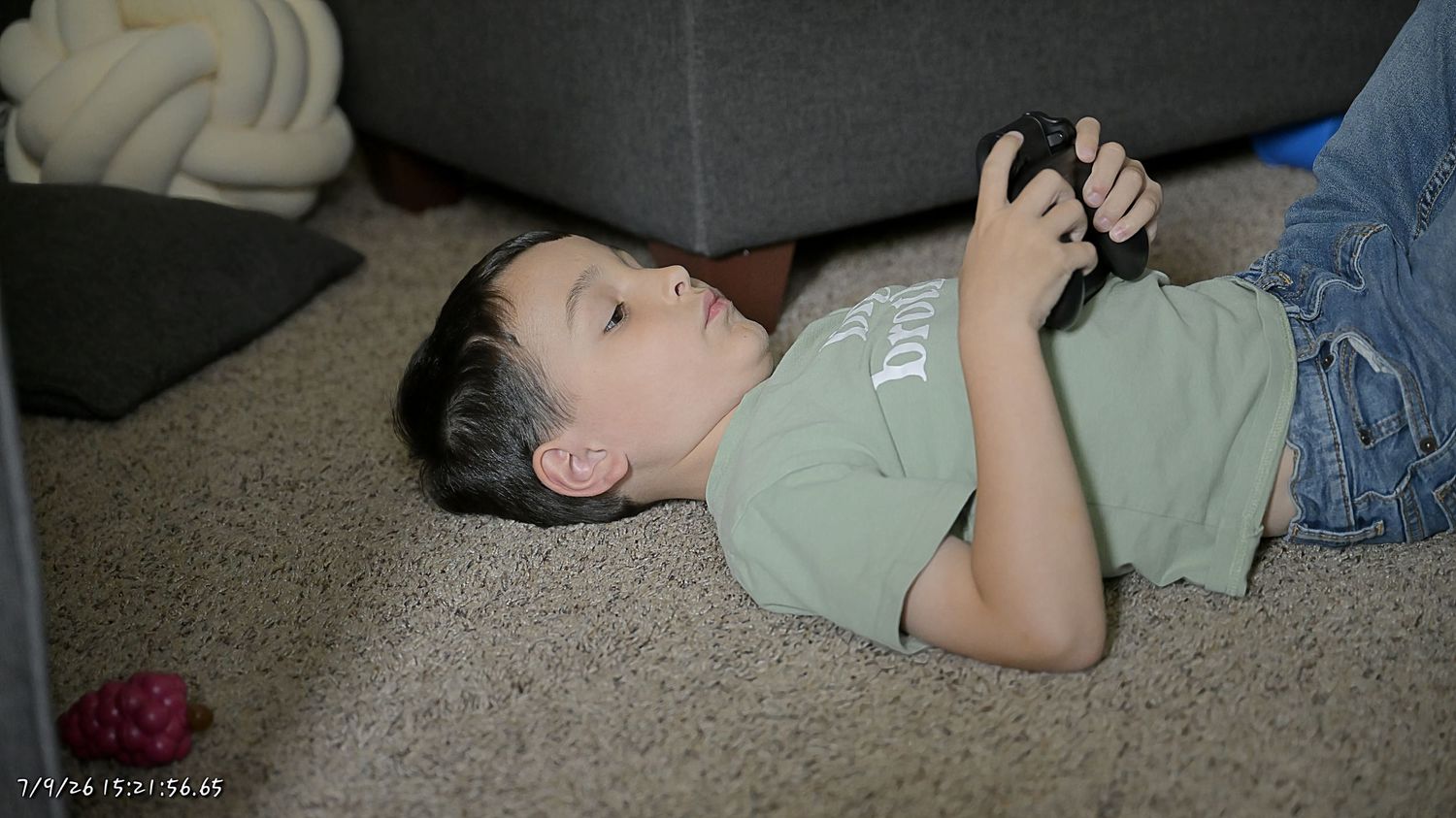 A young boy lying on carpet playing a handheld video game device in a dimly lit room.