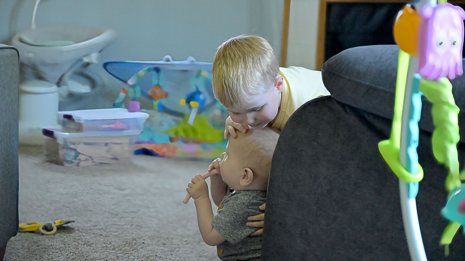 A toddler leans over to kiss a baby sitting on the floor near a gray couch.
