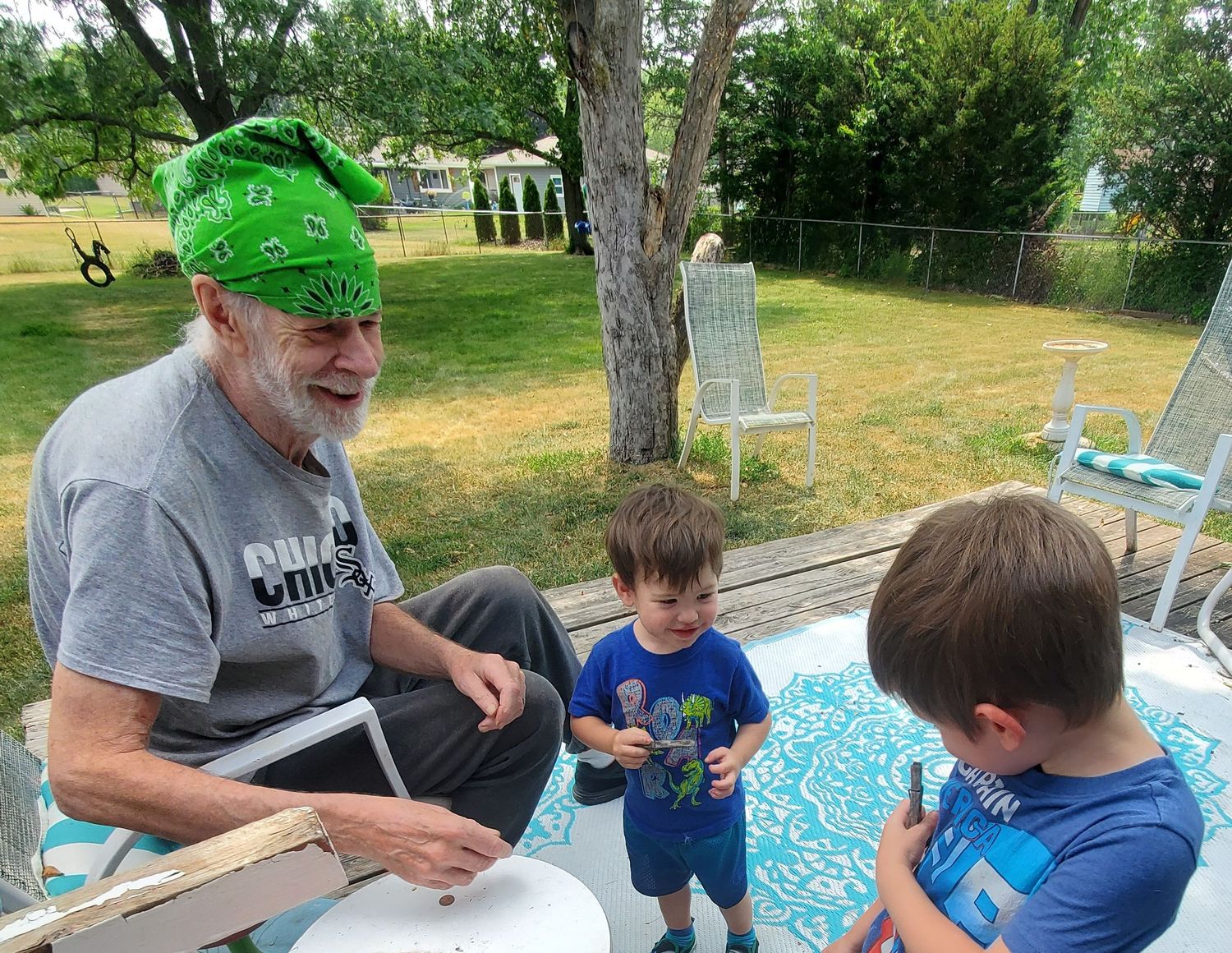 Older man in green bandana sits on porch interacting with two young boys in sunny backyard.