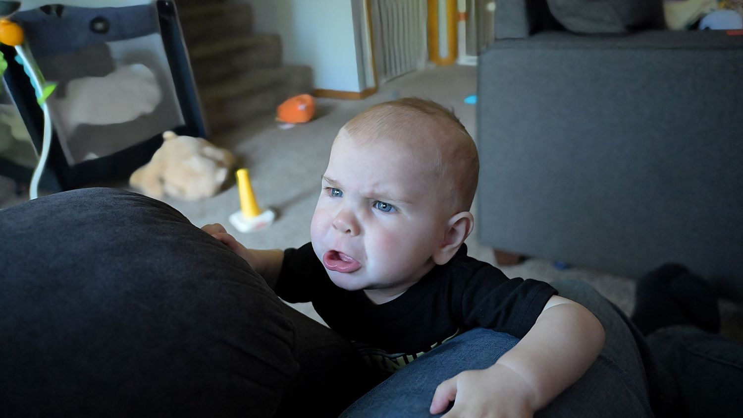 Fussy baby in black shirt leaning on gray sofa with toys in background.