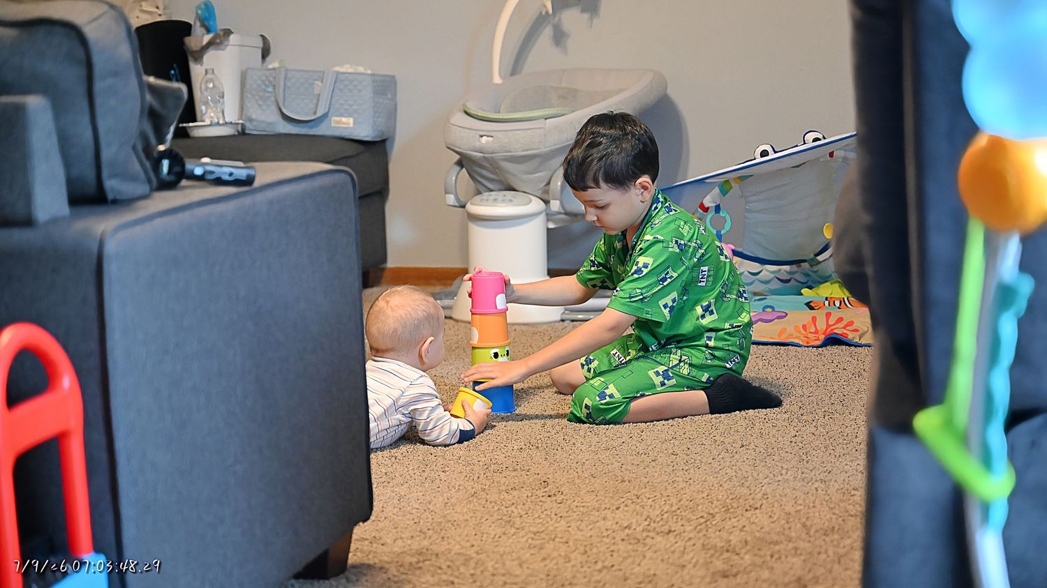 An older sibling in green pajamas playing with a baby on the floor with stacking toys.