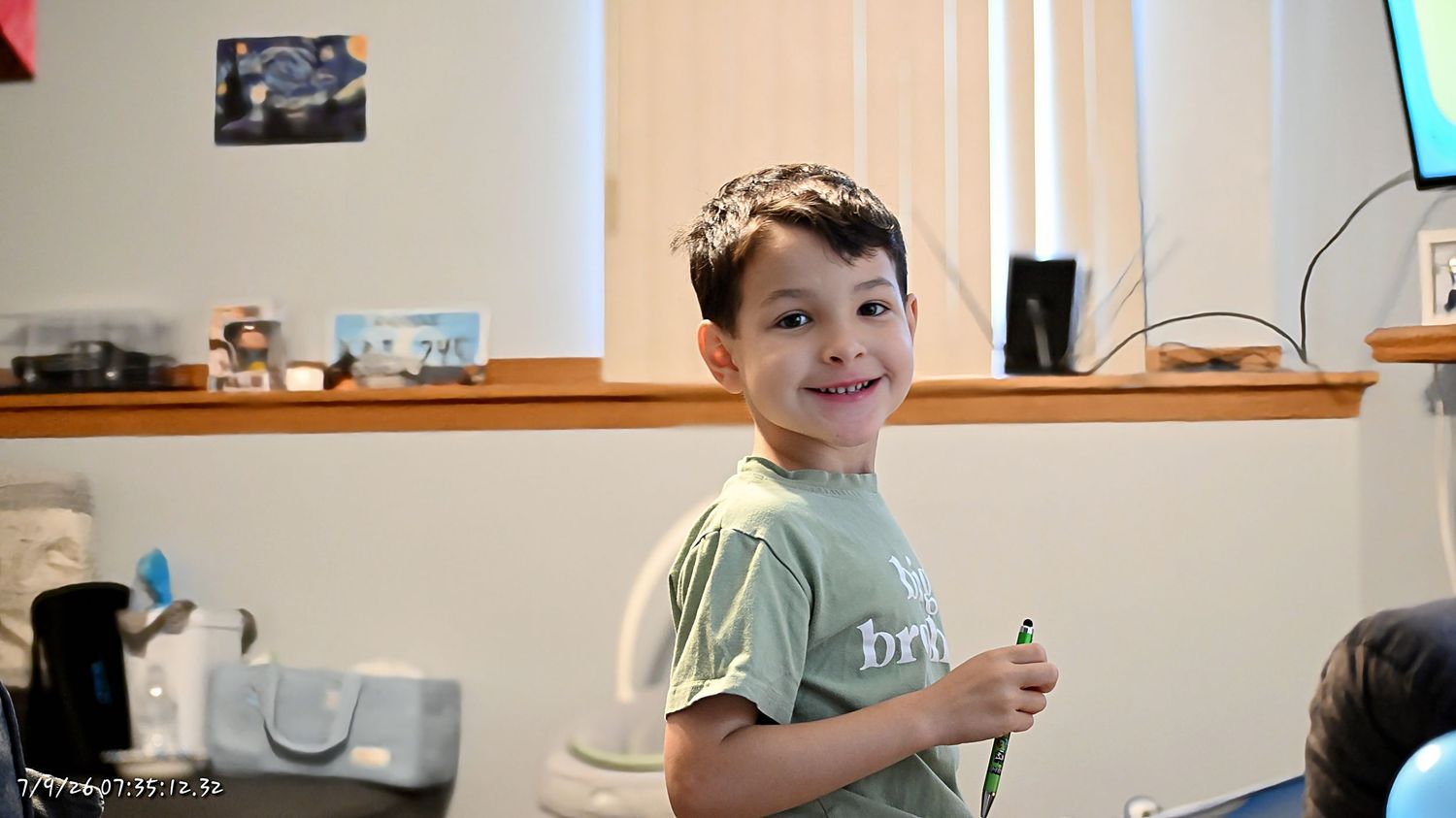 A smiling boy in a green t-shirt standing in a kitchen holding a small object.