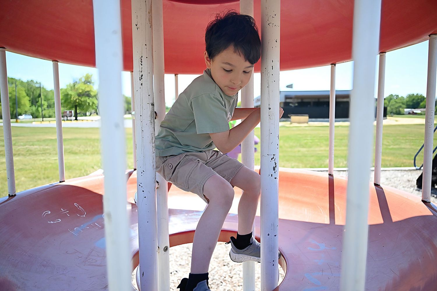 A young boy in a green shirt climbs inside a red and white merry-go-round at a sunny playground.