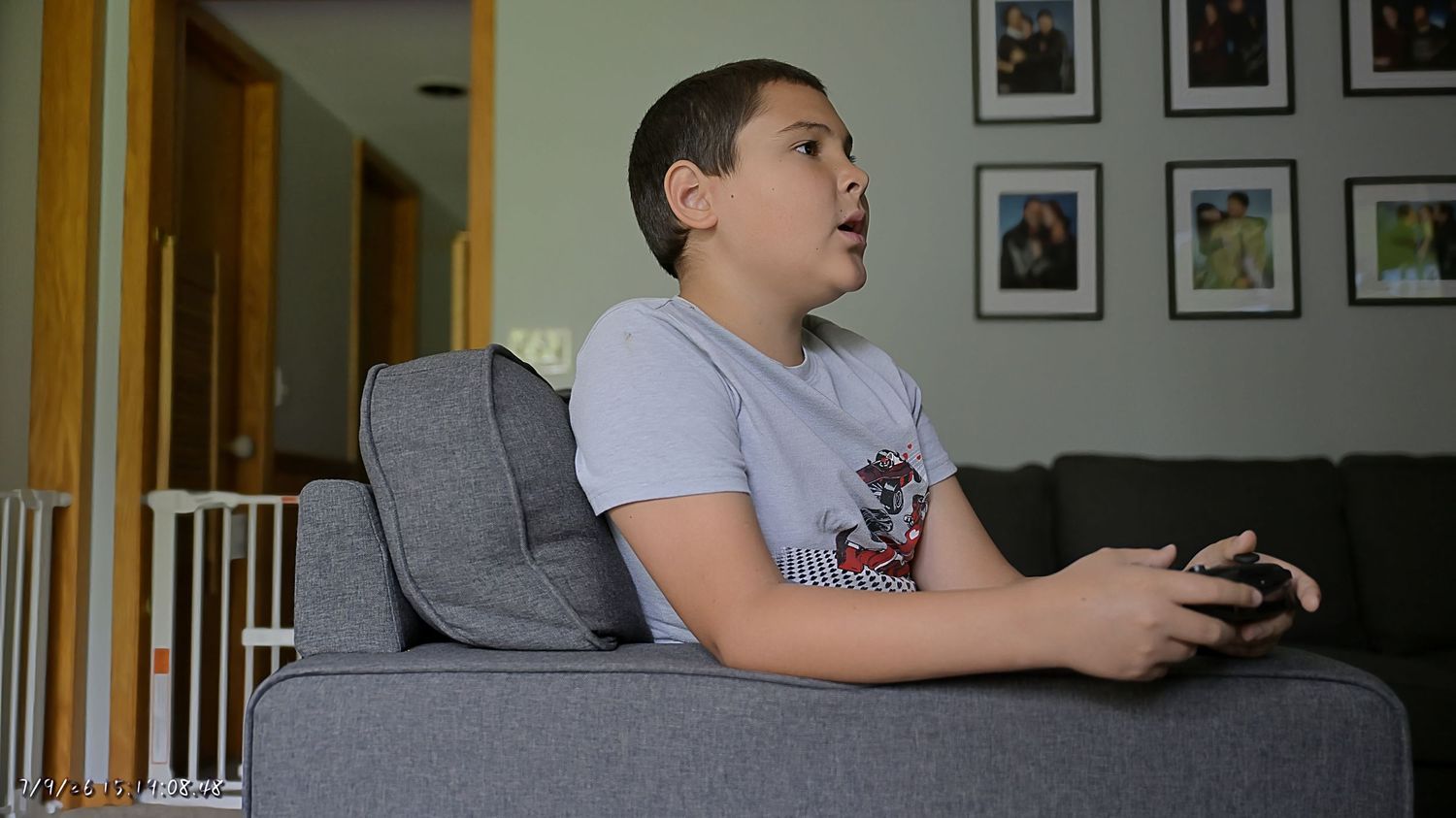 A young boy sitting on a gray sofa playing a video game with a controller in a living room.