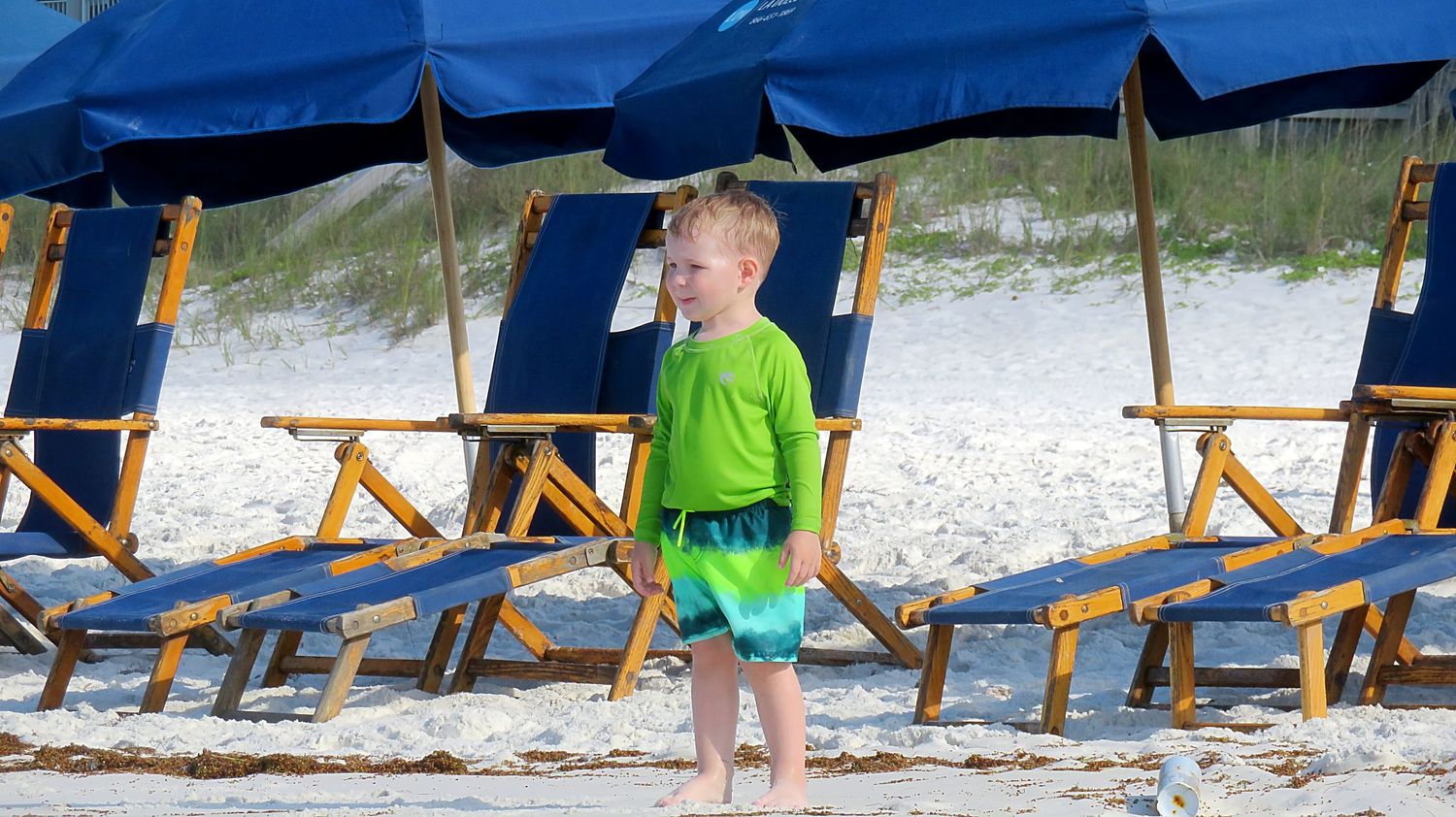 Young boy in green shirt stands among blue beach chairs and umbrellas on a sandy beach.