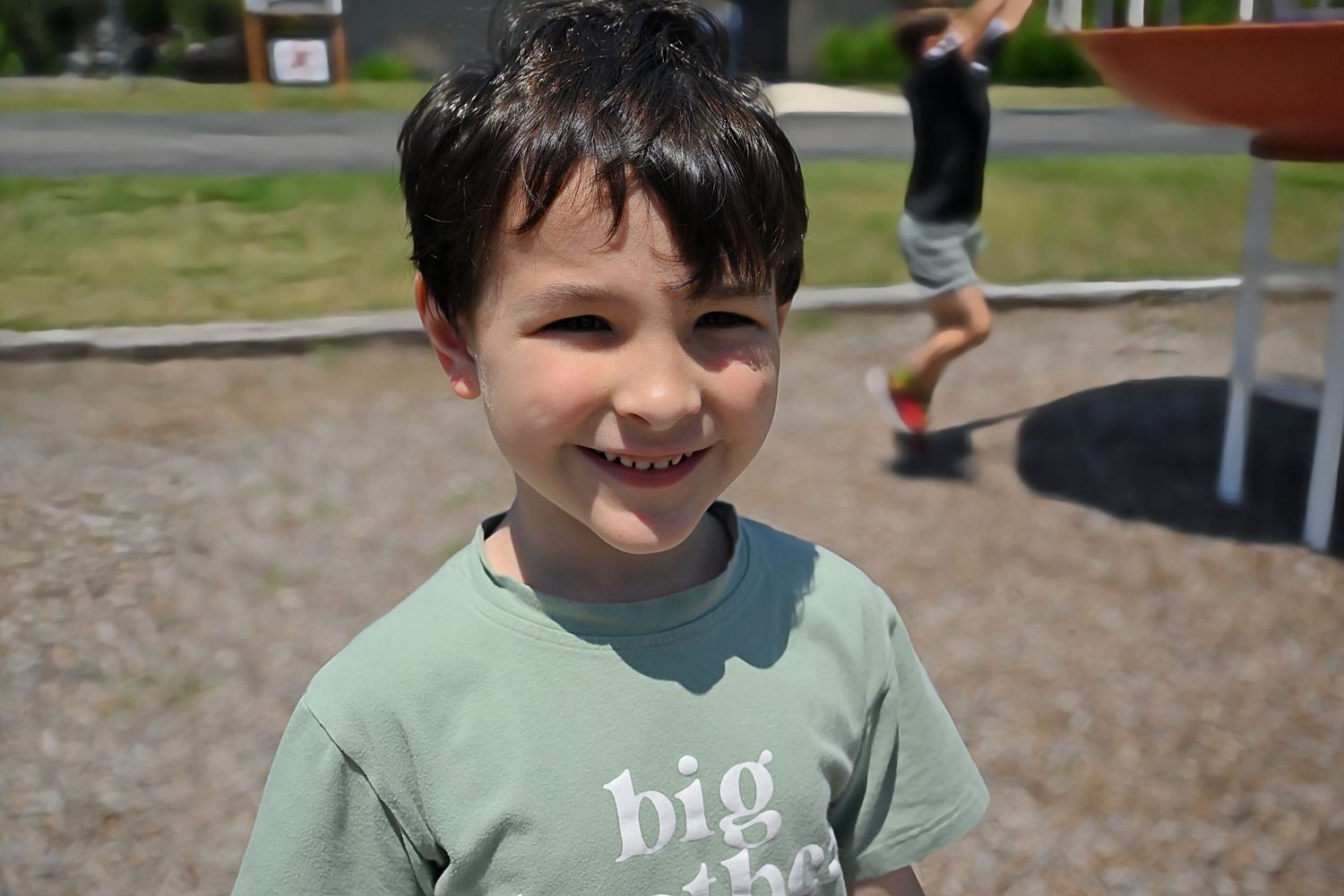 A cheerful young boy in a green big brother t-shirt smiles at the camera at a sunny playground.