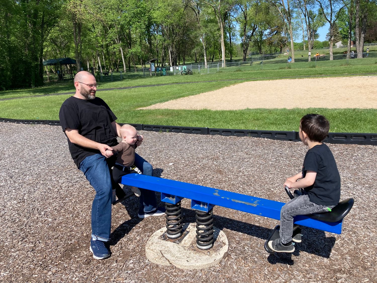 A father helps his young child on a blue spring seesaw at a sunny park playground.