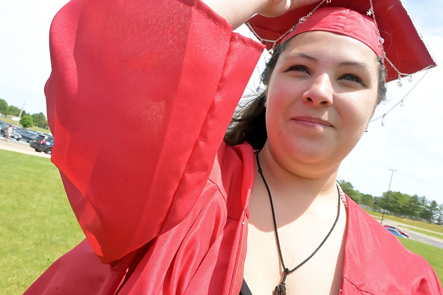 Young woman in red graduation cap and gown smiling outdoors on a sunny day.