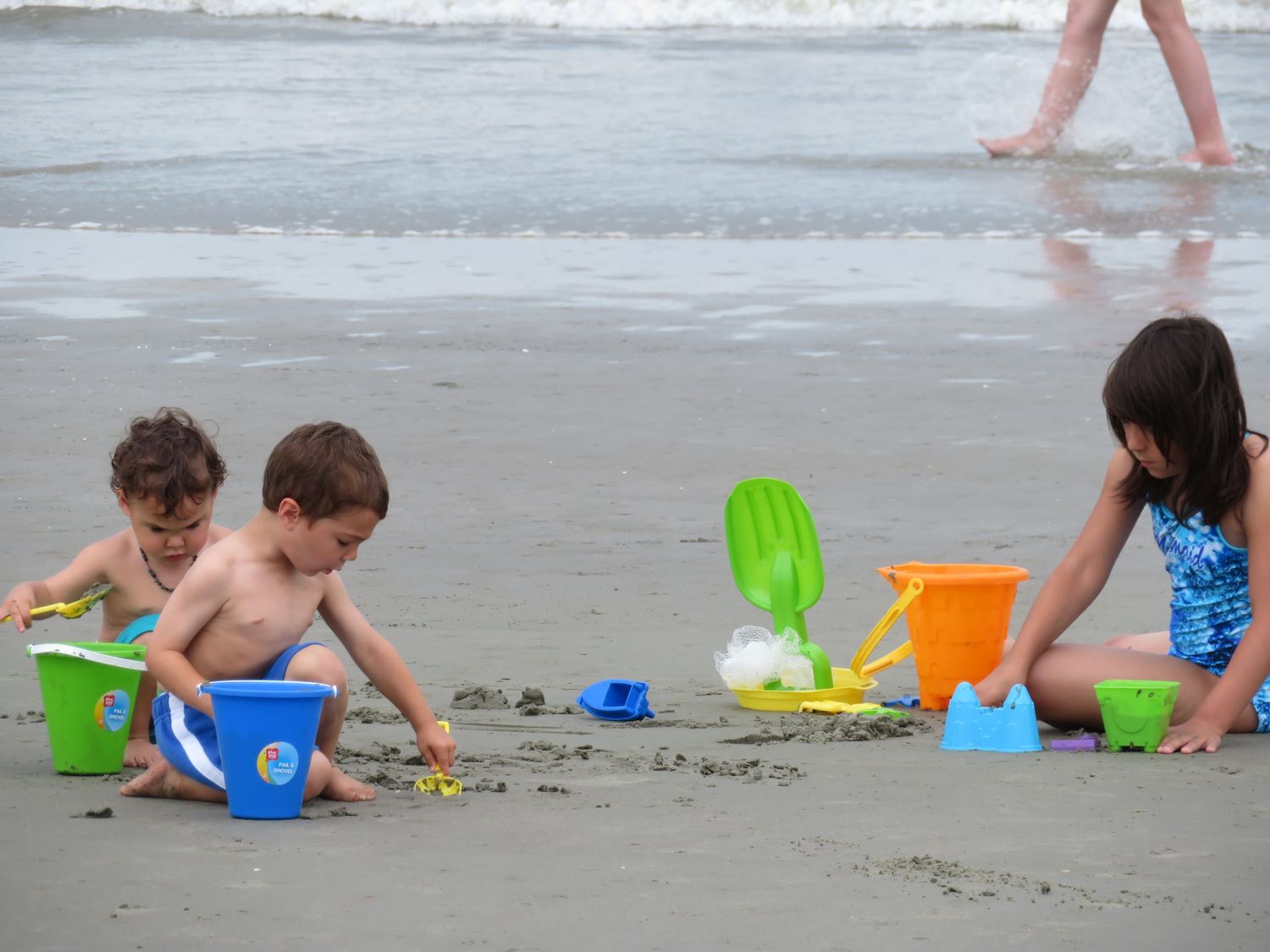 Three young children playing with colorful sand buckets and toys on a beach.