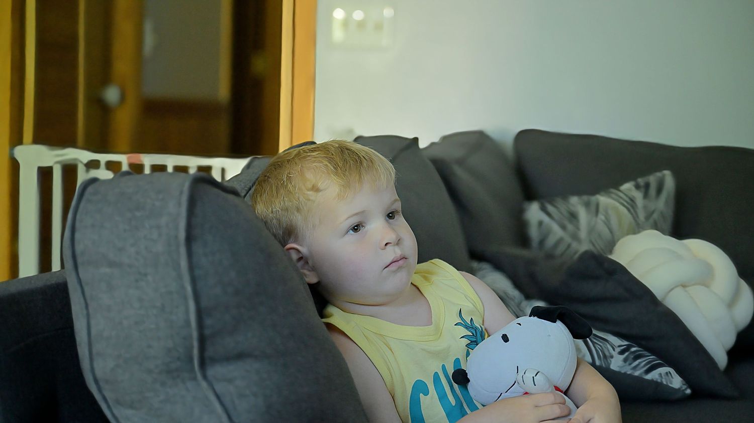 Toddler in yellow shirt relaxing on gray sofa holding a stuffed animal toy.