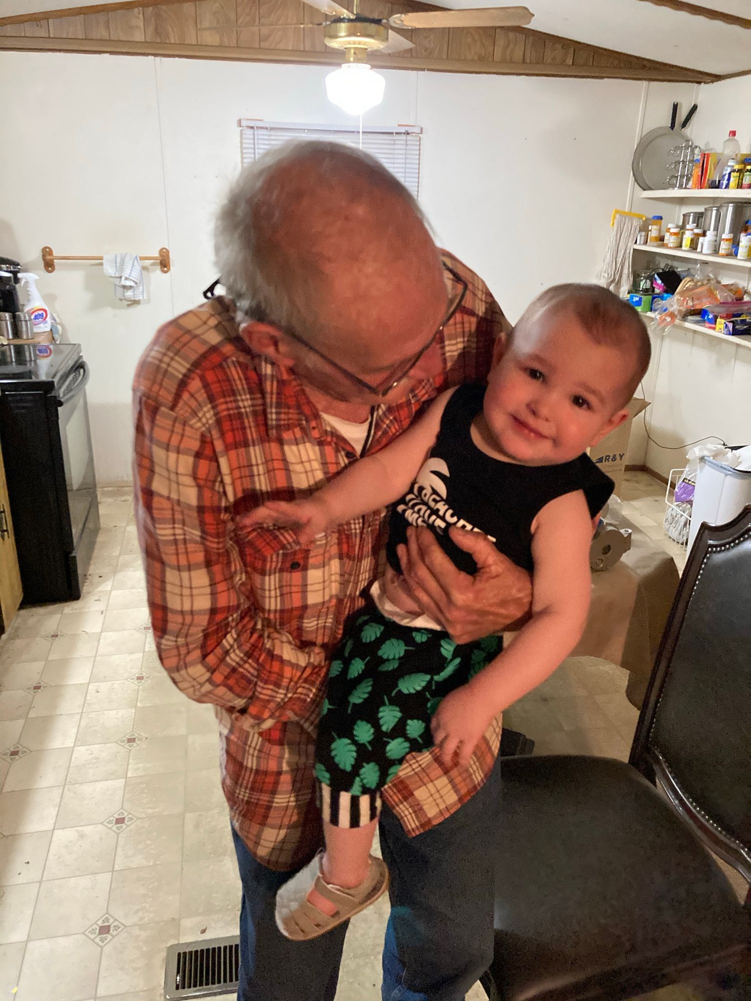 Elderly grandfather in plaid shirt holding smiling baby in black shirt indoors.