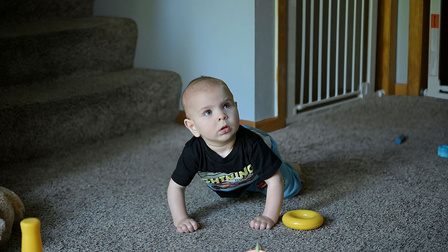 Baby in black shirt crawling on carpet near stairs with colorful toys nearby.
