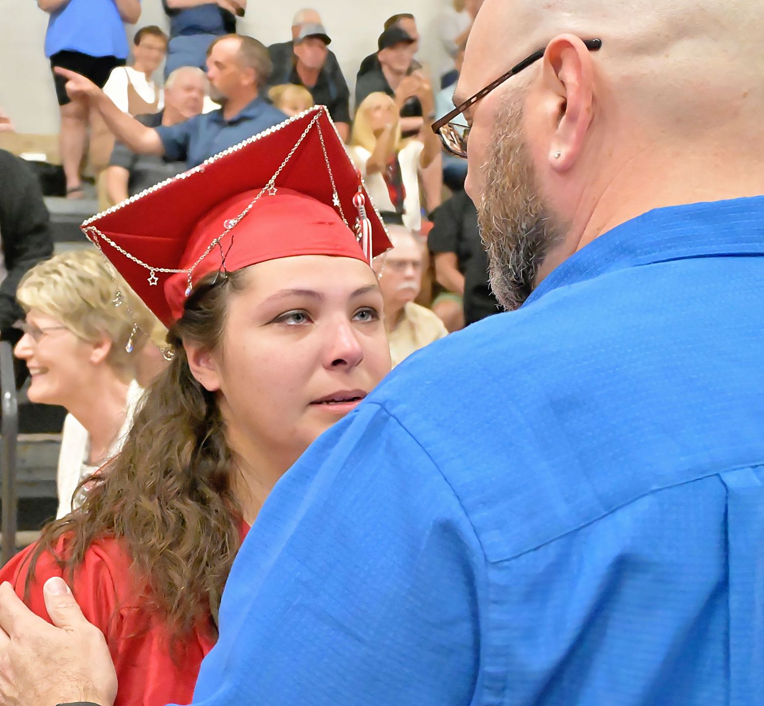 Emotional female graduate in red cap and gown shares a heartfelt moment with a man at graduation ceremony.