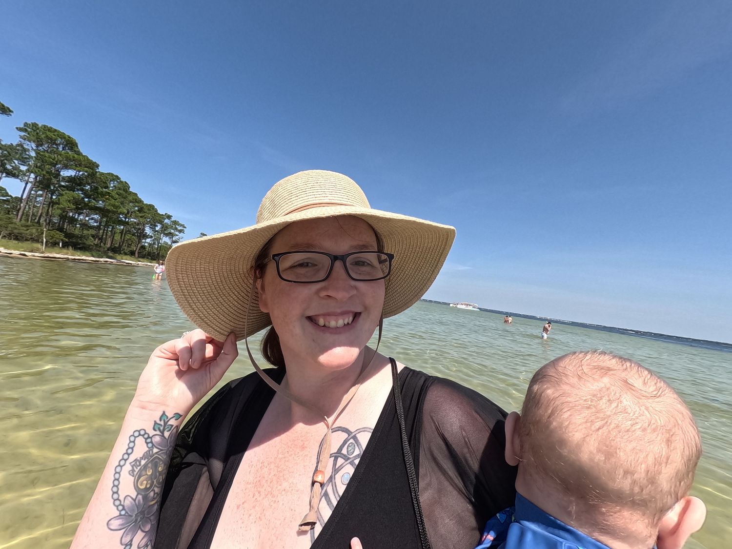 Smiling woman in wide-brim straw hat holding a baby at a sunny beach location.