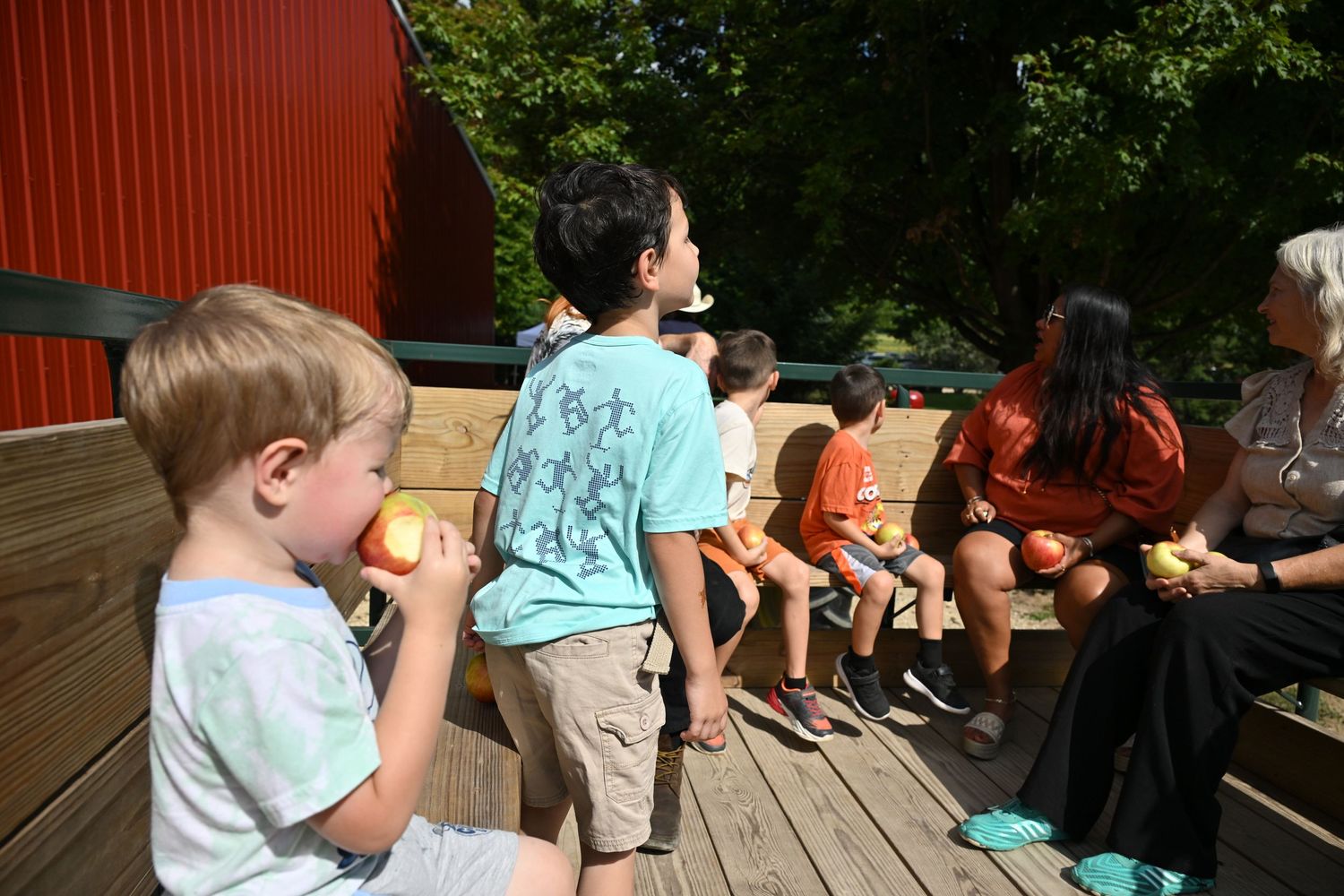 Children and adults sit on outdoor hay ride eating fresh apples on a sunny day.