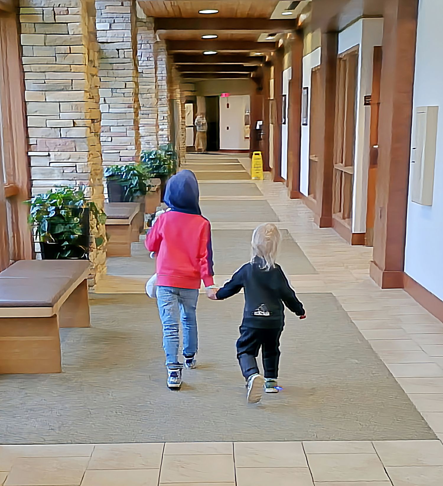 Two young children holding hands walk down a wide indoor corridor with wooden columns.