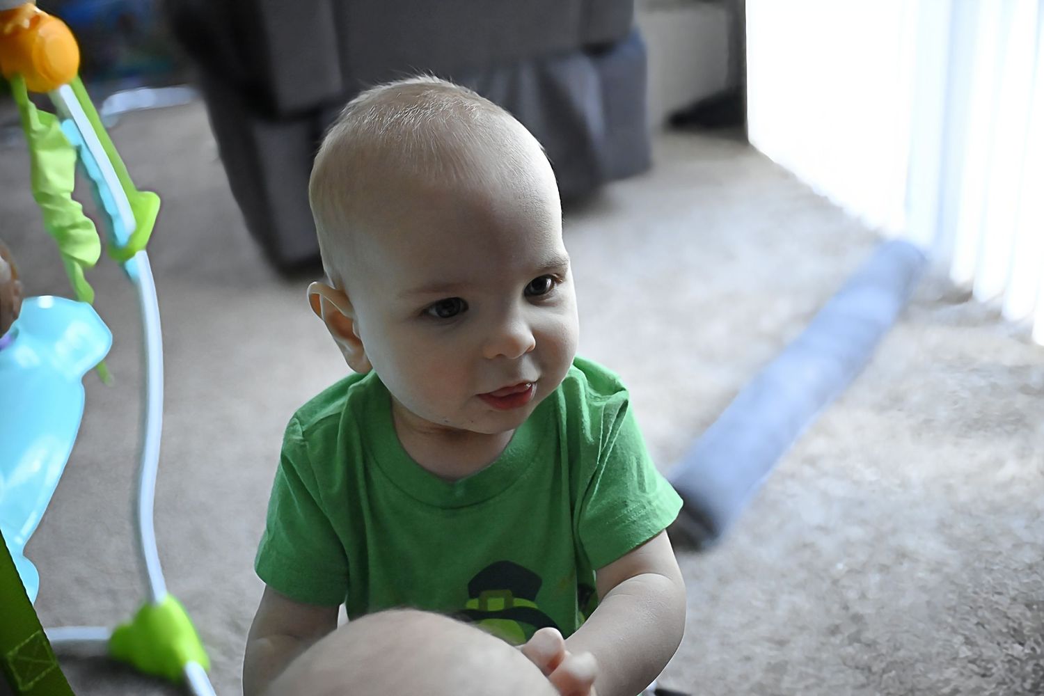 An adorable baby in a green shirt sitting near a colorful activity toy on carpet.