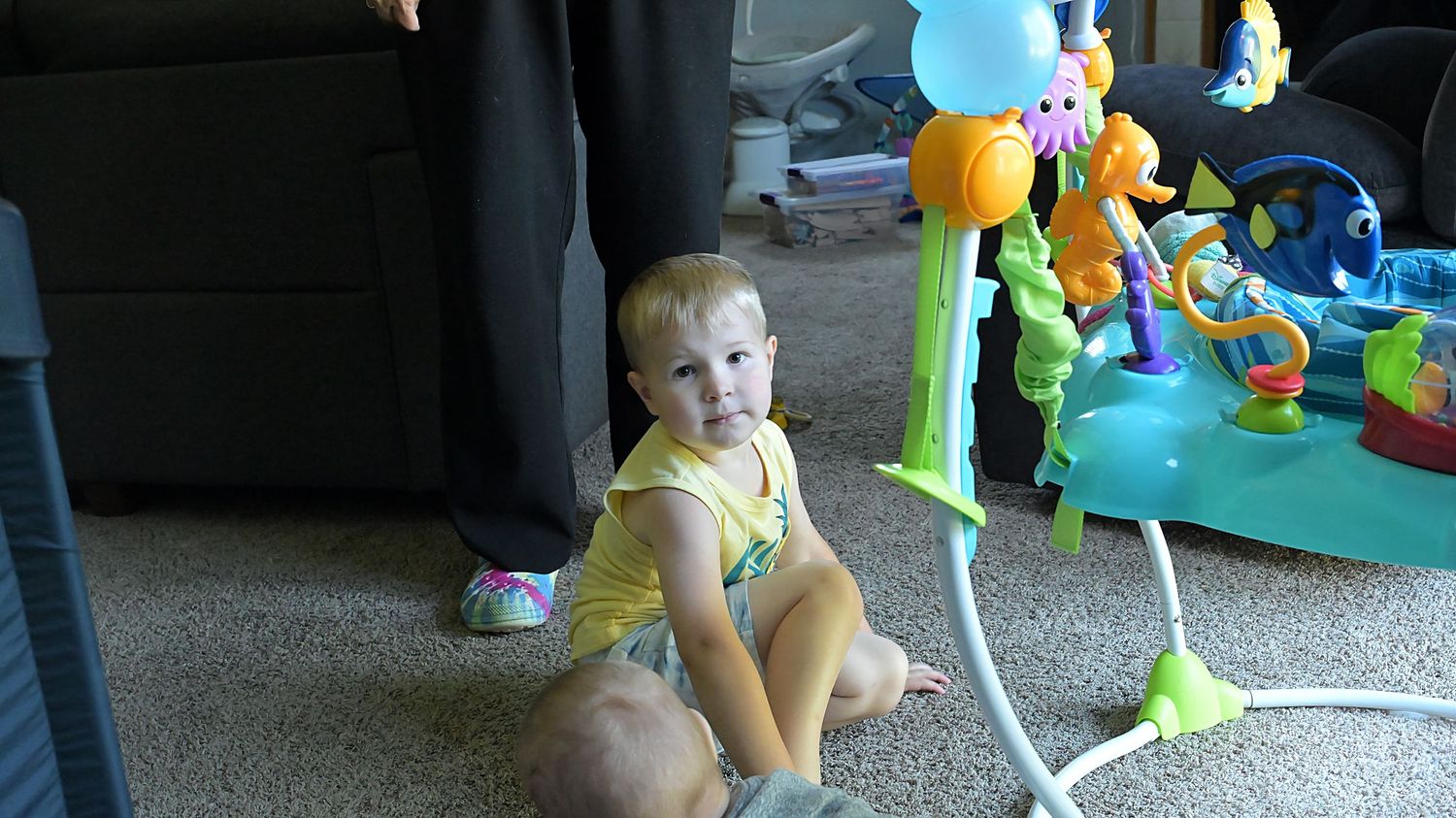 A young toddler sits on carpet near a colorful baby activity jumper toy indoors.