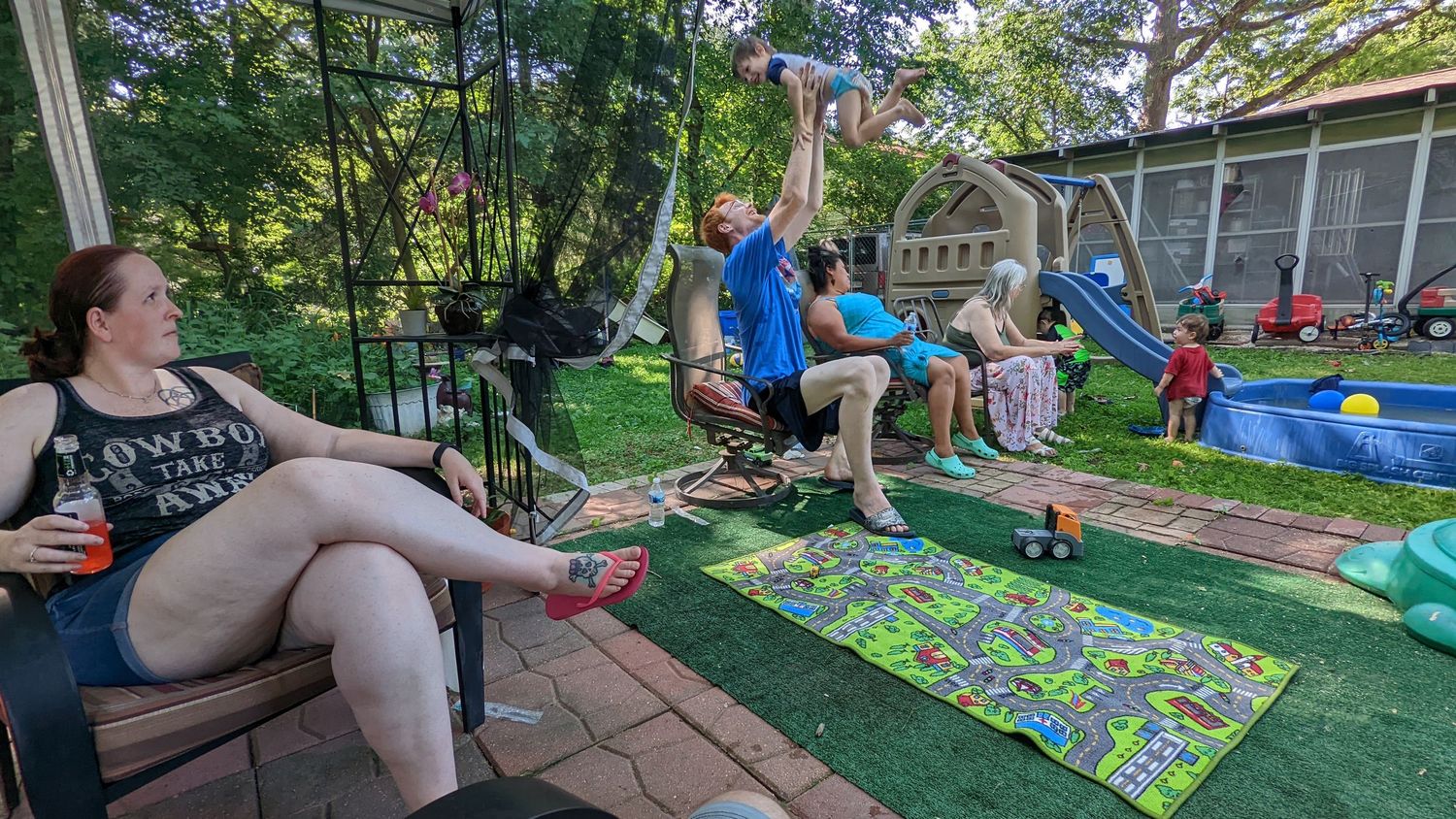 Family relaxing on backyard patio while children play in yard on summer day.